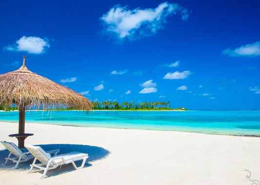 Image of an idyllic beach scene with blue skies, aqua water and white sand. There are a couple of beach chairs and shaded spot on the left hand side